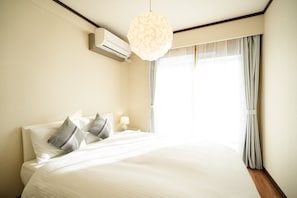 Quarto de hotel com uma cama grande, uma mesa de cabeceira com luminária, um aparelho de ar condicionado e uma janela com cortinas.