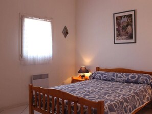 A bedroom with a bed, a bedside table with a lamp, a window with a curtain, and a wall-mounted picture. - Spacious 3-Room Villa with Mezzanine, Terrace & BBQ, Near Grazel Beach - Sleeps 6 (Gruissan)