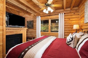A log cabin bedroom with a bed, a television, a fireplace, and a window with curtains. - Rise n' Shine by Beyond Expectations: Cozy Upscale Cabin w/ Fireplace & Hot Tub (Sevierville)
