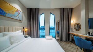 A hotel room with a large bed, a desk, a chair, a TV, and a view of a cityscape through the windows. - La Beaute Hotel Quy Nhon (Quy Nhon)