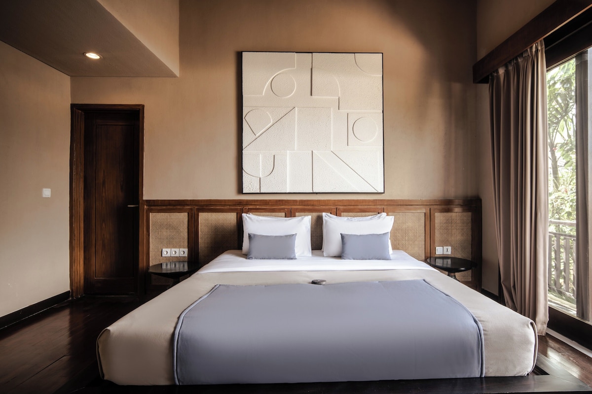 A large bed with a headboard, a wall-mounted artwork, a window with curtains, and a door.