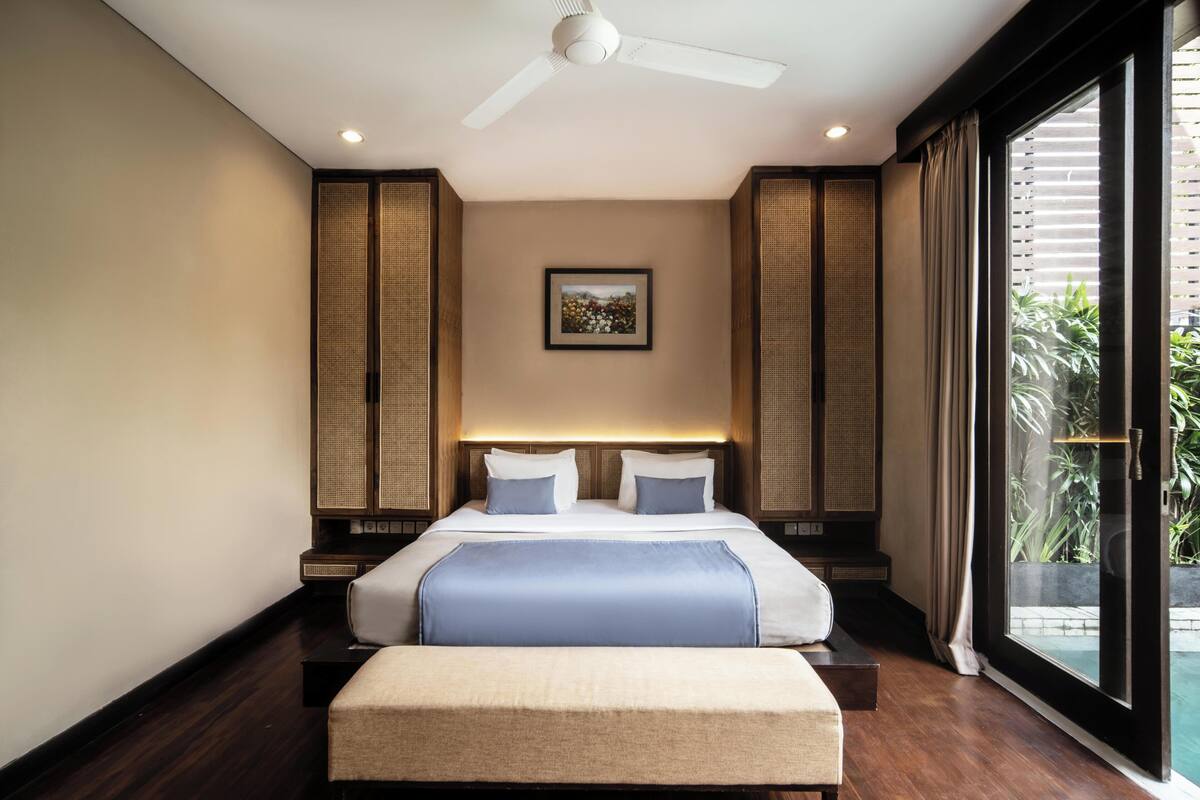 Une chambre d’hôtel moderne avec un grand lit, un ventilateur de plafond, un tableau encadré au mur et une vue sur la verdure.