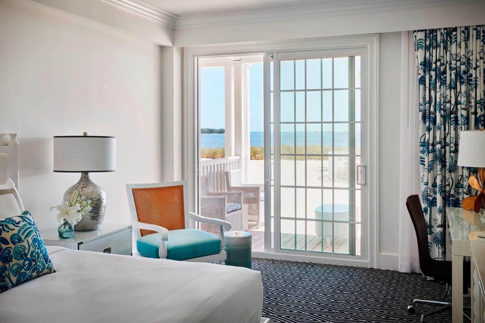 A hotel room with a bed, a chair, a table, a lamp, and a view of the ocean through a sliding glass door.