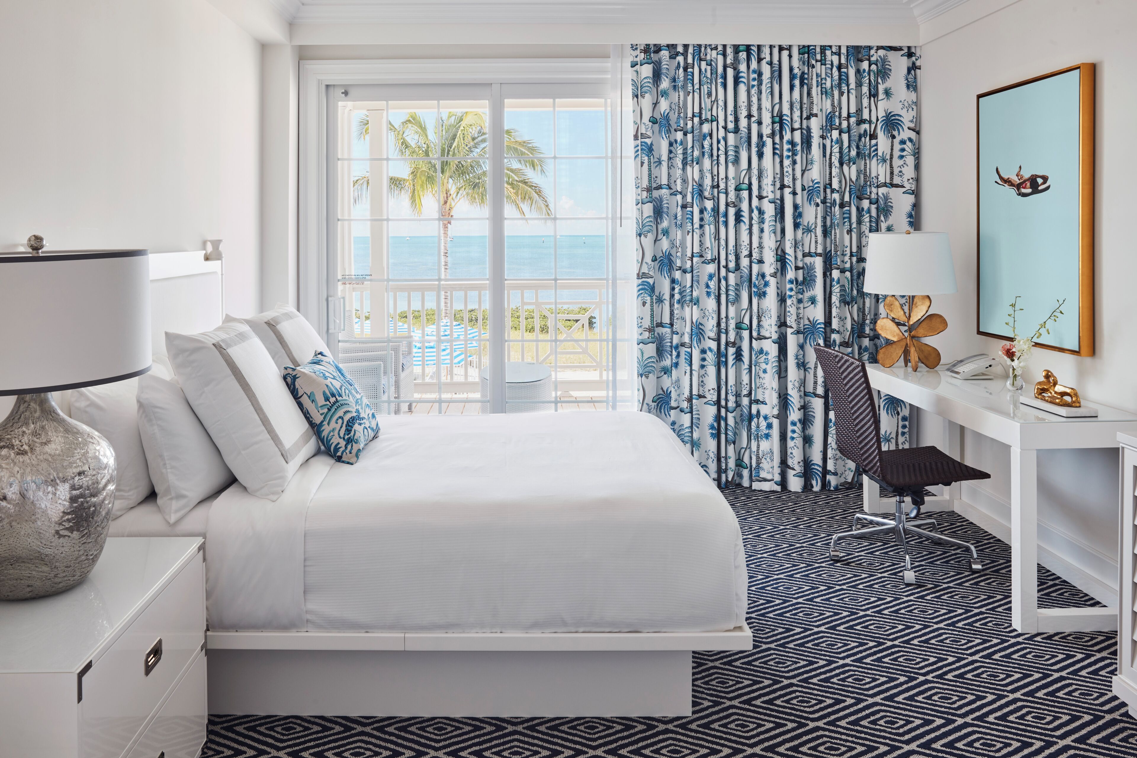 A hotel room with a bed, a desk, a chair, a lamp, a large window with a view of the ocean, and a tropical-themed curtain.