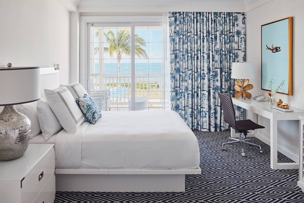 Une chambre d’hôtel comprenant un lit, un bureau, une chaise, une lampe, une grande fenêtre donnant sur l’océan et un rideau aux motifs tropicaux.