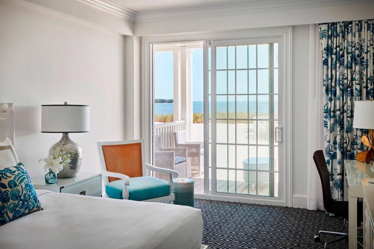 Une chambre d’hôtel avec un lit, une chaise, une table, une lampe et une vue sur l’océan, visible à travers une porte-fenêtre coulissante.