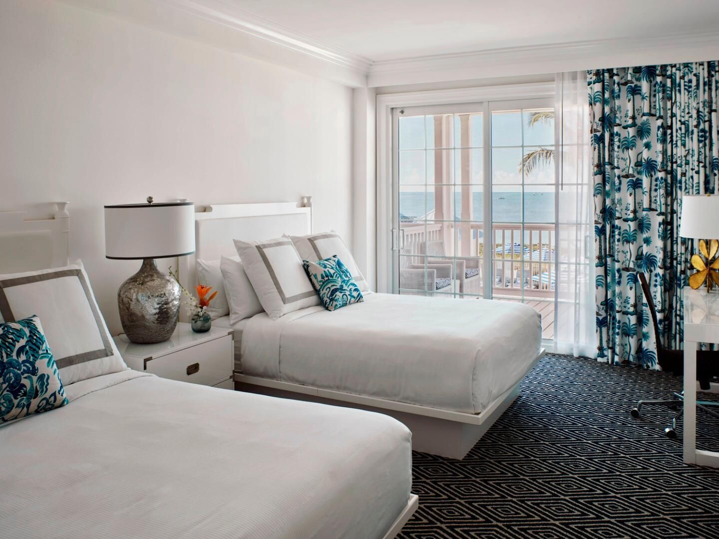 A hotel room with two beds, a large window with a view, a patterned carpet, and a nightstand with a lamp.