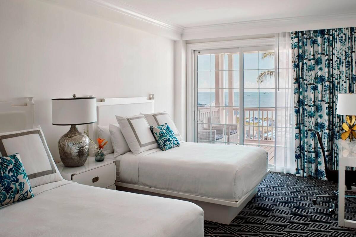 Une chambre d’hôtel avec deux lits, une grande fenêtre donnant sur un beau paysage, une moquette à motifs et une table de chevet avec une lampe.