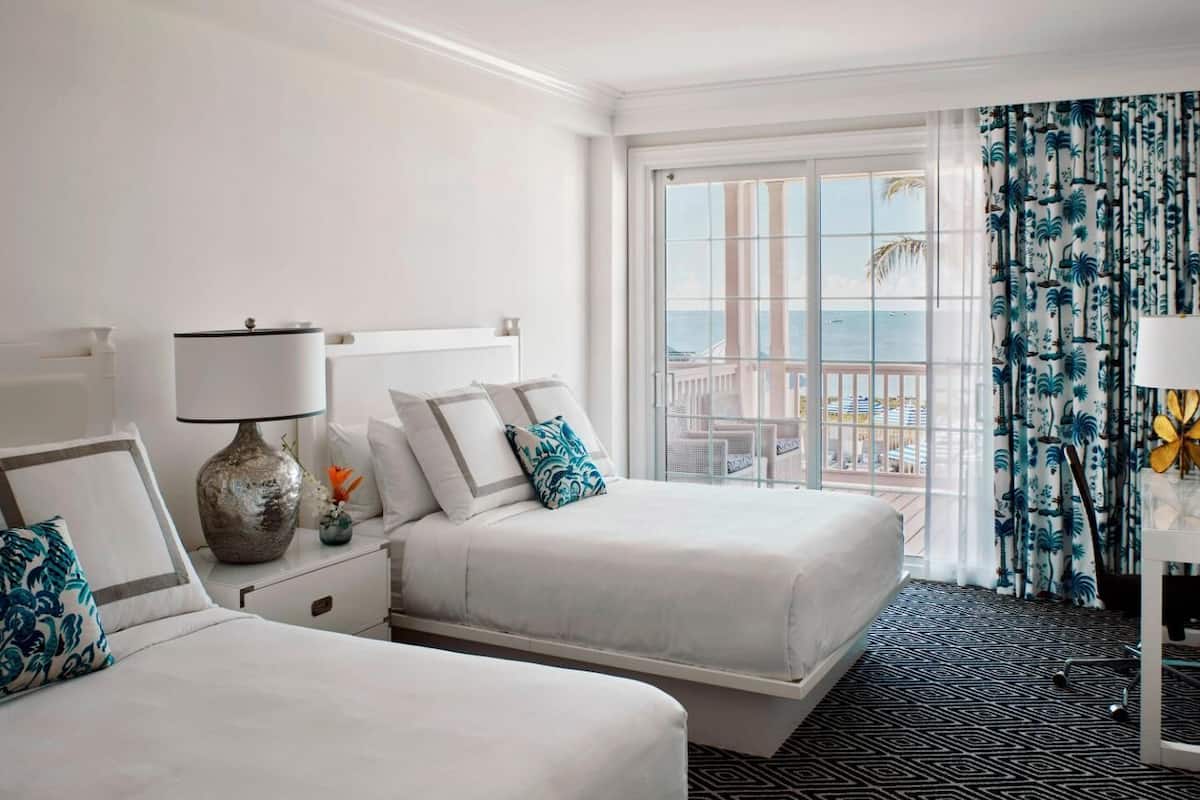 A hotel room with two beds, a large window with a view, a patterned carpet, and a nightstand with a lamp.