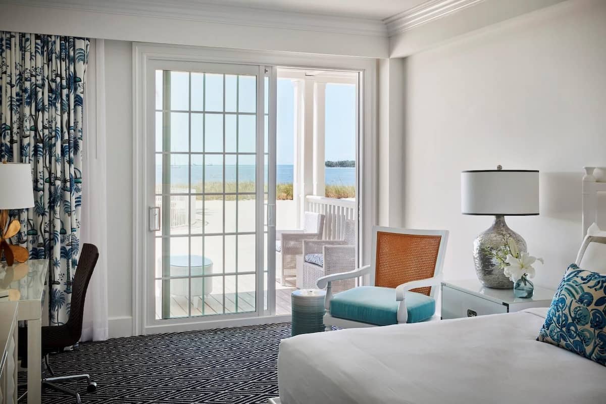 A hotel room with a bed, a desk, a chair, a lamp, and a view of the beach.