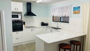 Cocina compacta con armarios blancos, extractor de aire negro, microondas, tostadora y fregadero. Hay dos taburetes de madera en la encimera.
