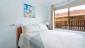 Un dormitorio con cama, ventana que ofrece vista a un edificio de ladrillo y un cuadro con temática de playa.