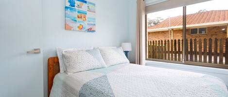 A bedroom with a bed, a window with a view of a brick building, and a wall with a beach-themed artwork.