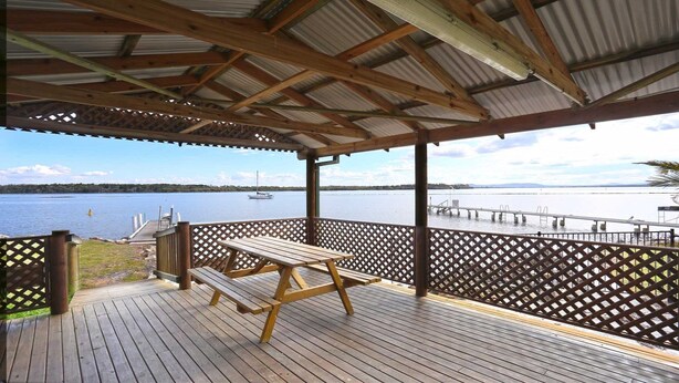 A covered deck with a wooden table and benches, overlooking a calm body of water with a dock and a boat.