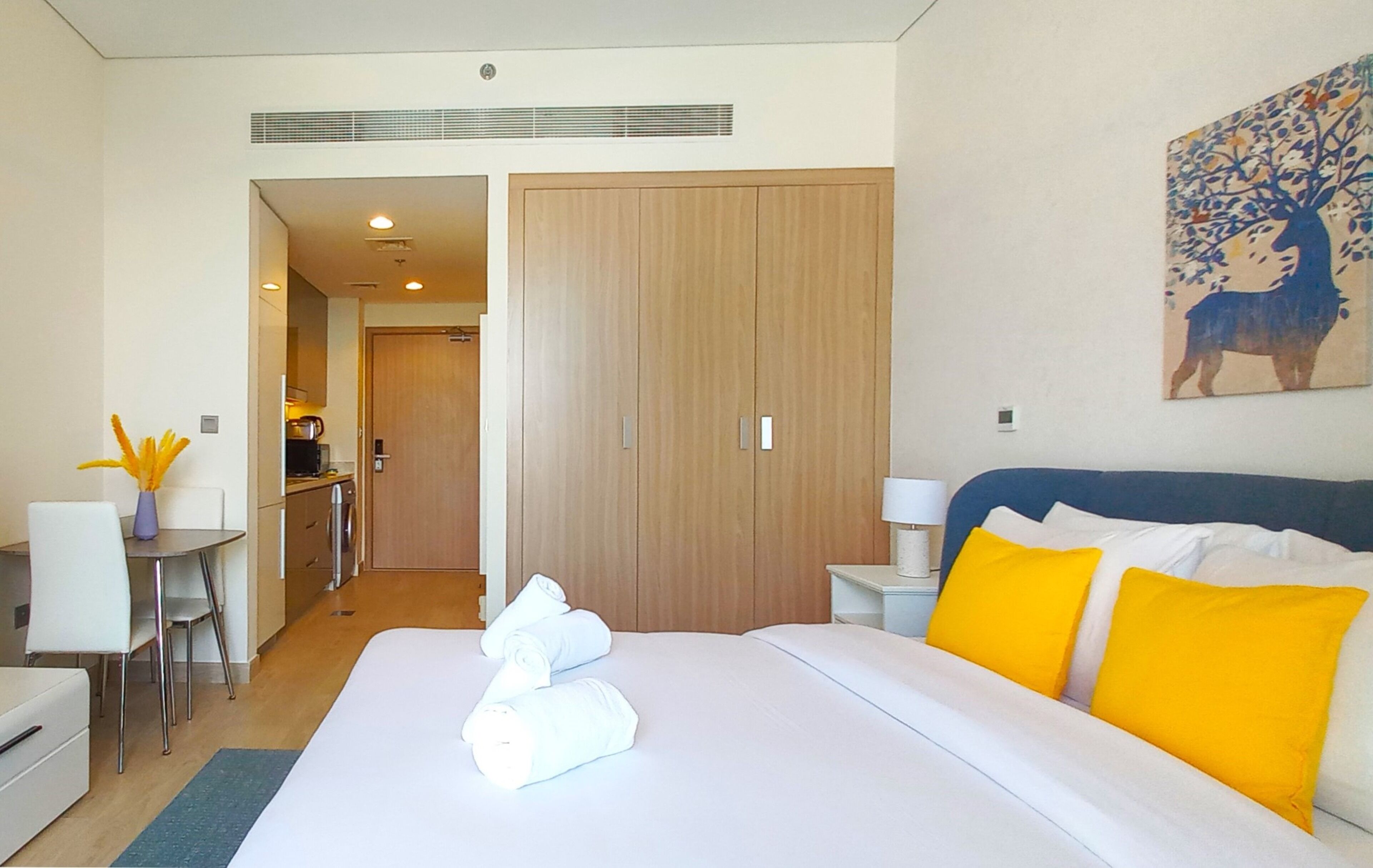 Una habitación de hotel con cama, escritorio, silla, una mesita con un florero y un cuadro en la pared.
