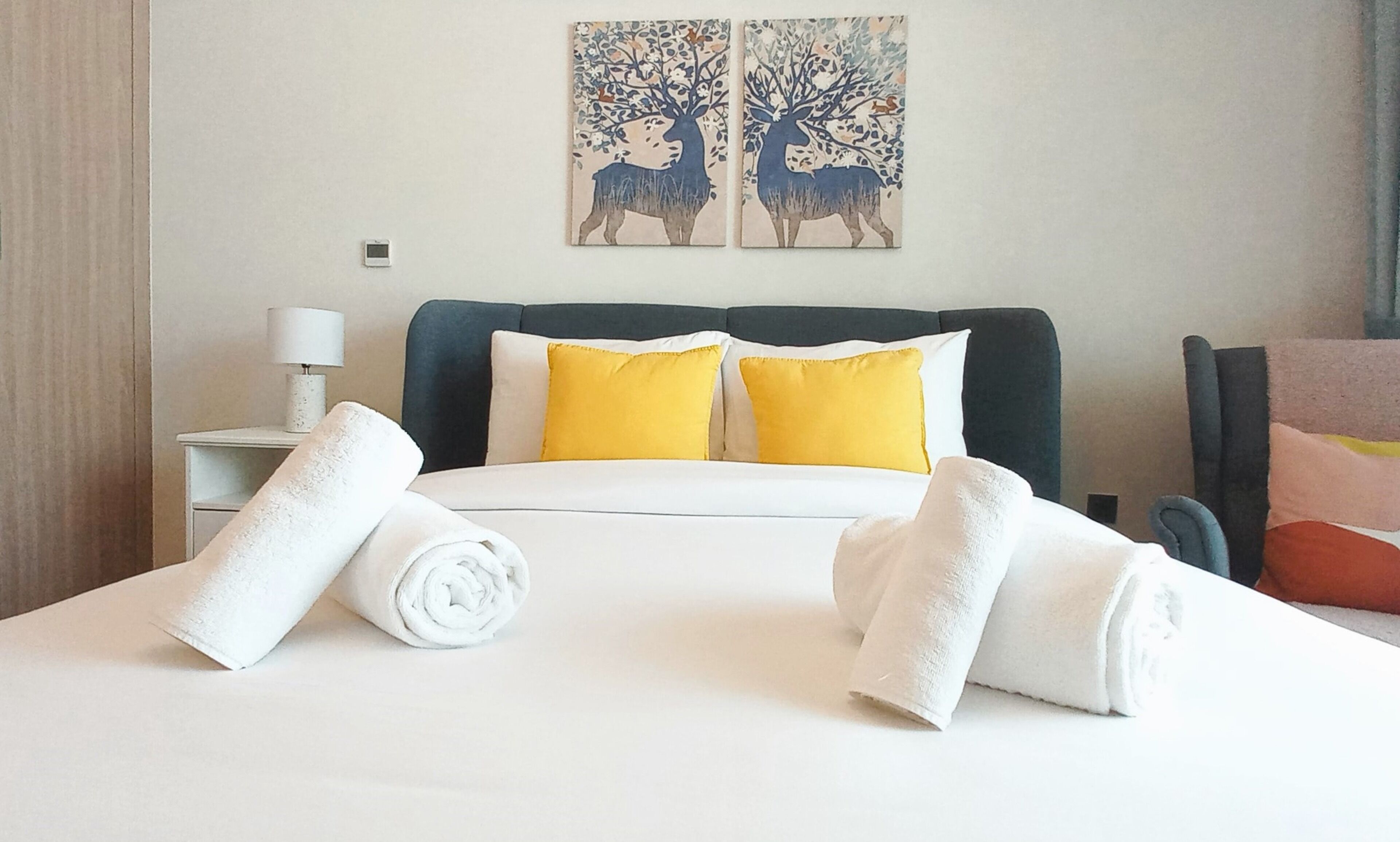 A hotel room with a bed, two yellow pillows, a white headboard, and two rolled towels on the bed.