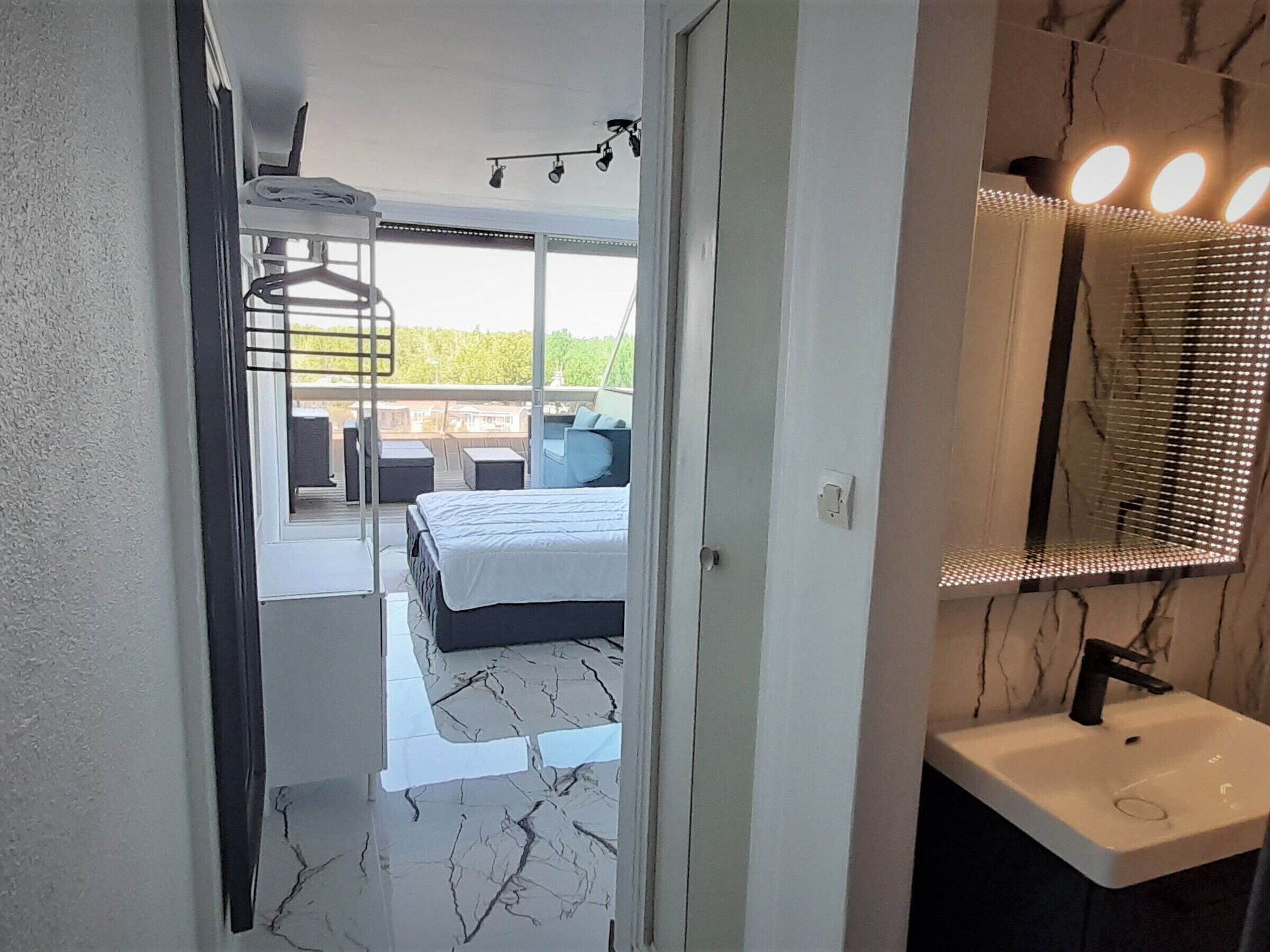 A bedroom with a bed, a balcony with seating, a bathroom with a sink and mirror, and a view of greenery outside.