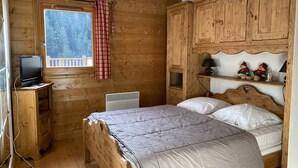 A wooden cabin room with a bed, a TV on a wooden stand, a window with a view, and wooden cabinets.