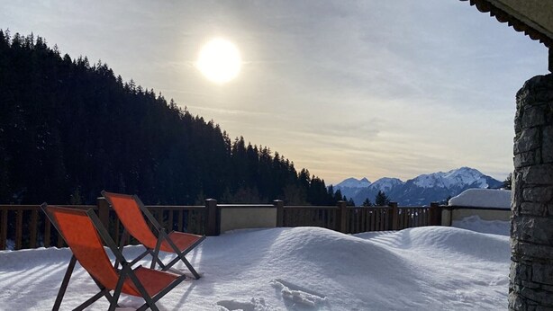 A snow-covered deck with two orange chairs, a stone wall, and a mountain view.