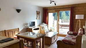 A cozy room with a wooden table, a sofa, a TV, and a sliding glass door leading to an outdoor deck.