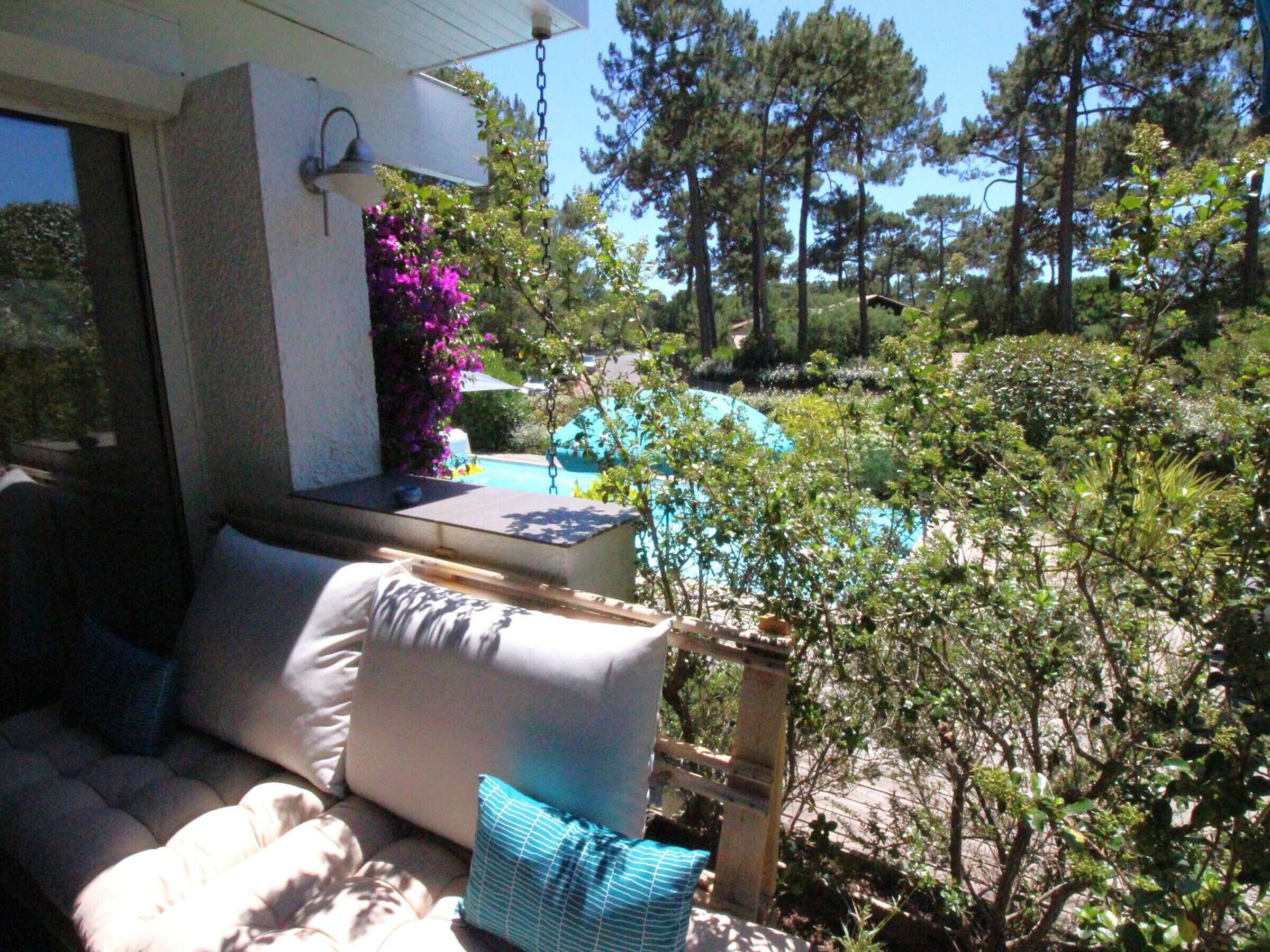 Un patio con un sofá y cojines, una mesita con una lámpara, y vista a una piscina rodeada de árboles y arbustos.
