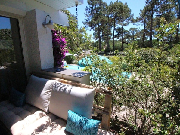 A patio with a sofa and cushions, a small table with a lamp, and a view of a pool surrounded by trees and shrubs.