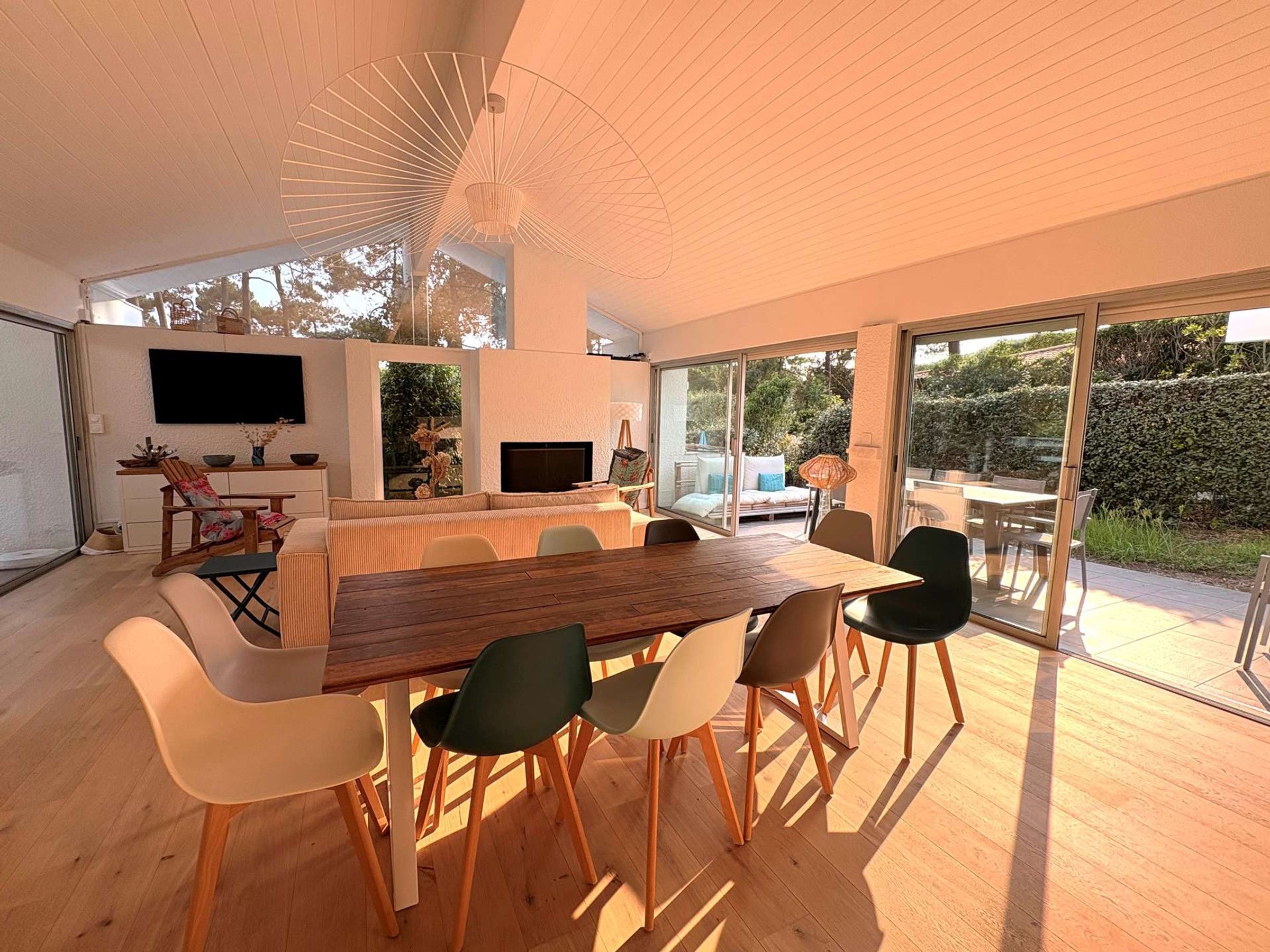 A modern dining area with a large wooden table and chairs, a flat-screen TV mounted on the wall, and a sliding glass door leading to an outdoor patio.