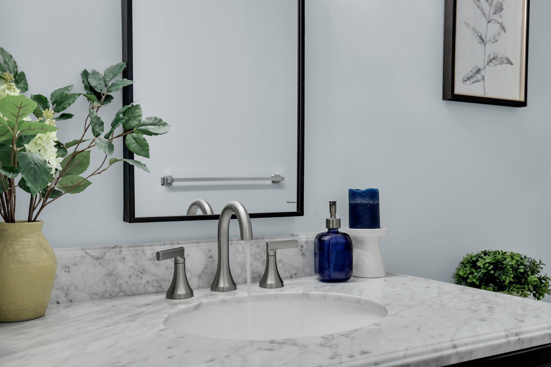 Ein Badezimmer mit einem weißen Marmorwaschbecken, zwei Wasserhähnen, einem Handtuchhalter, einem blauen Seifenspender und einem gerahmten Pflanzenprint an der Wand.
