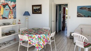 A dining area with a colorful tablecloth, white chairs, and a painting on the wall. - Apartment with balcony, garden, and beach access (Jullouville)