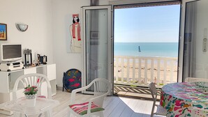 A balcony with a view of the beach, a table with a colorful tablecloth, and a white TV on a stand. - Apartment with balcony, garden, and beach access (Jullouville)