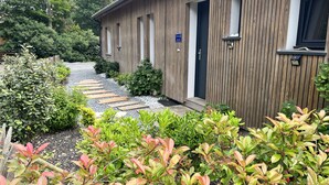 A wooden building with a door and windows, surrounded by greenery and a gravel path.
