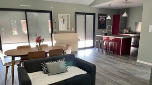 A modern living room with a black sofa, a dining table with chairs, and a kitchen area with red cabinets and a stainless steel refrigerator.