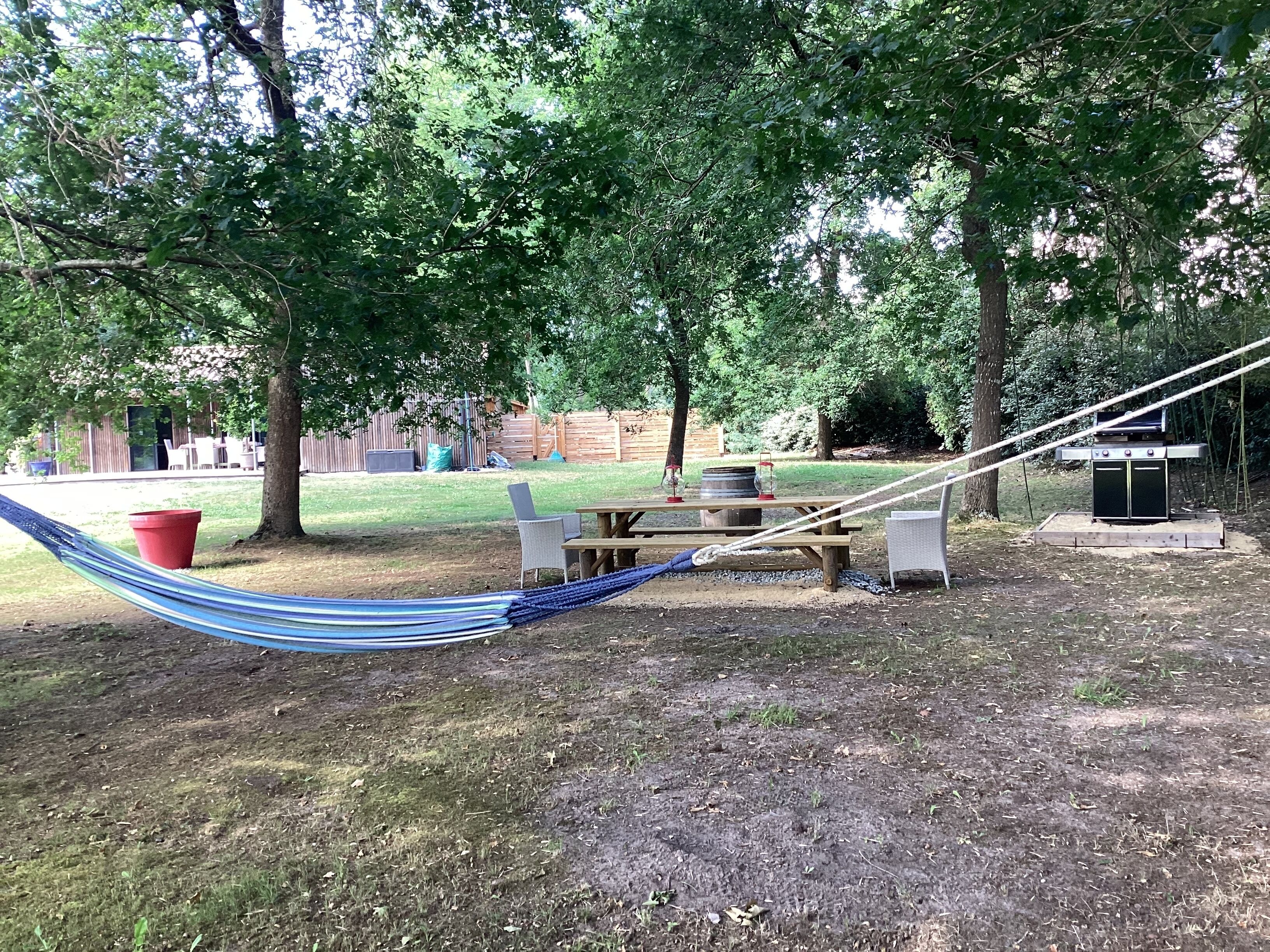 Una hamaca, una mesa de picnic y una parrilla en un patio trasero con árboles y una valla de madera.