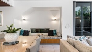 A modern living room with a wooden dining table, a built-in bench with cushions, a large potted plant, and a glass sliding door leading to a balcony. - Kokos 3 - Beach Houses of Byron (Byron Bay)