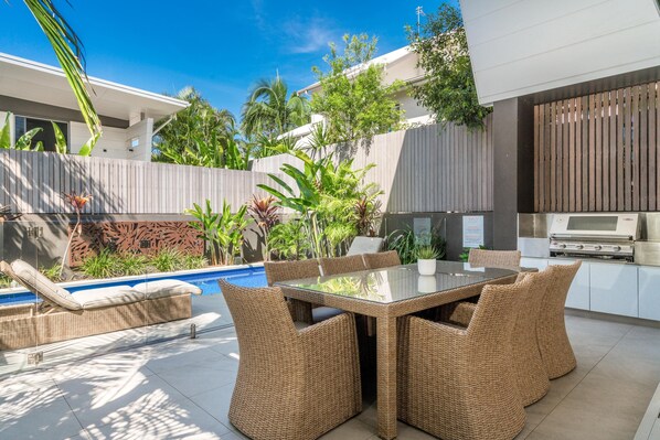 Eine moderne Außenpatio mit einem Tisch aus Glasplatten und Korbstühlen, umgeben von üppiger Bepflanzung und einem Swimmingpool.