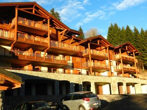 Un bâtiment en bois à plusieurs étages, doté de balcons, qui ressemble probablement à un hôtel ou à un chalet, avec une base en pierre et un espace de stationnement à l’avant.