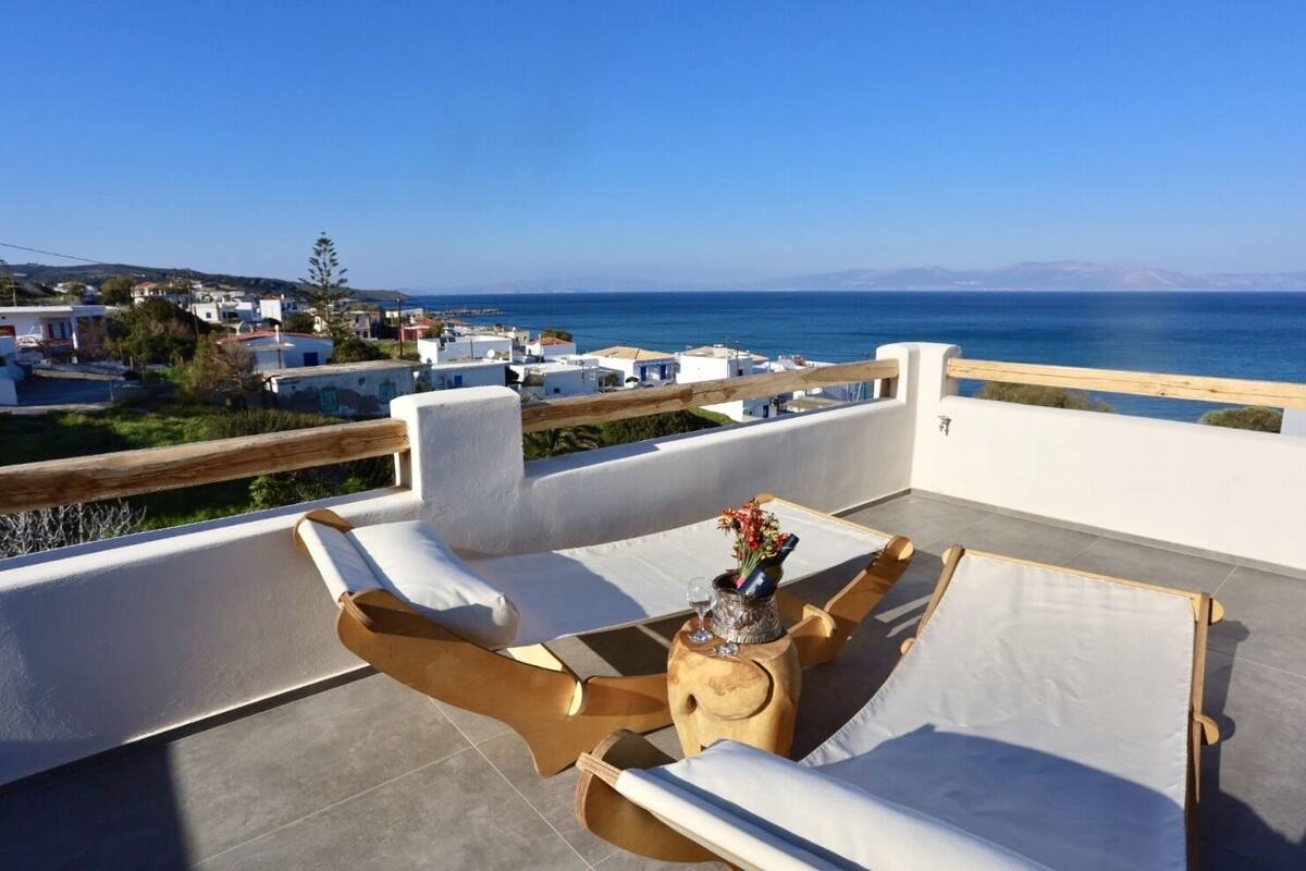 Terraço com assentos brancos acolchoados e uma mesa, com vista para uma cidade costeira e o mar.