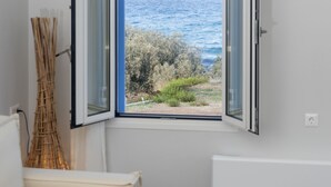 Une pièce avec un canapé blanc, une table ronde en bois, une fenêtre offrant une vue dégagée sur la mer et la verdure, et un radiateur.