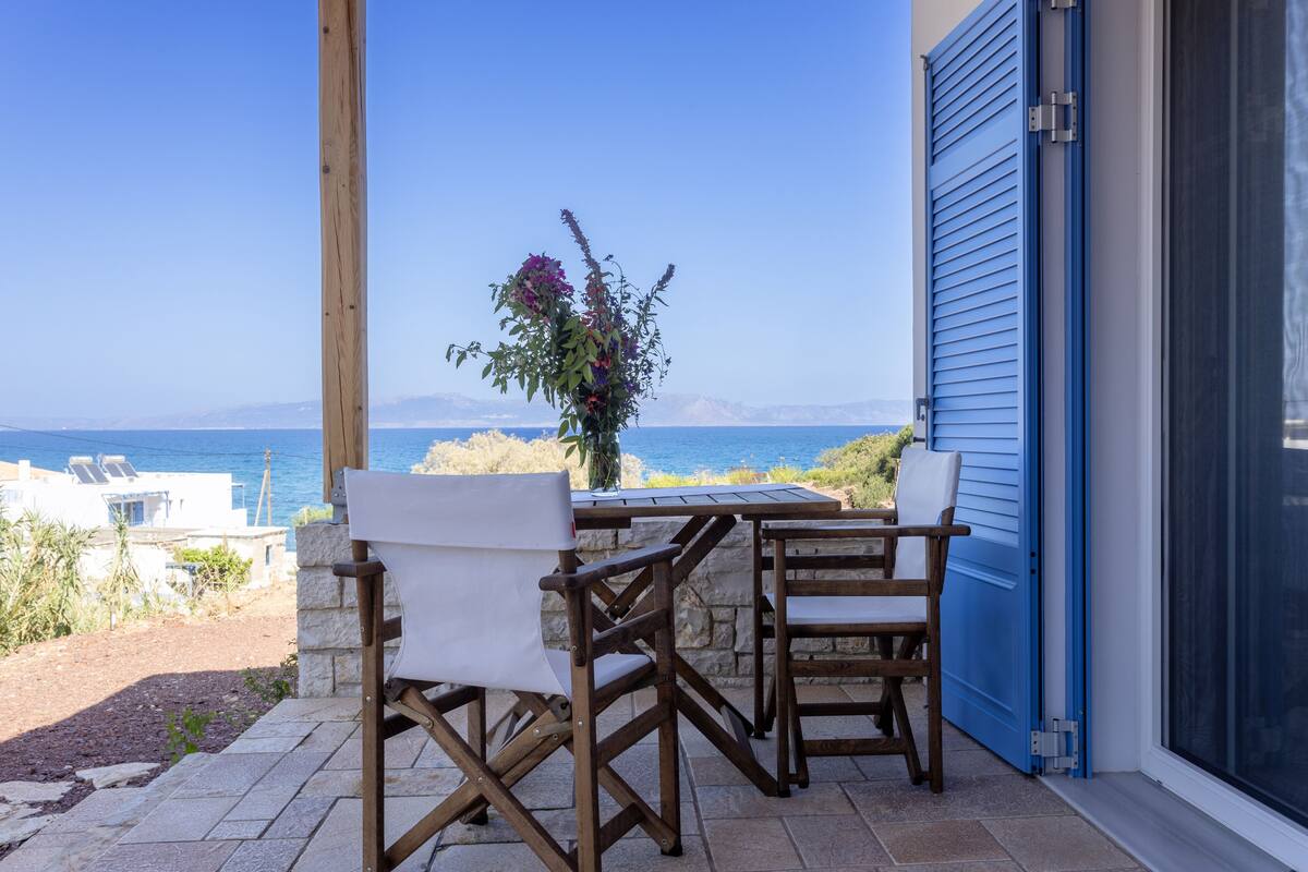 Um pátio com duas cadeiras de madeira e uma mesa, com vista para o mar e montanhas.