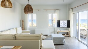 Un salon moderne avec un canapé, une table basse et un téléviseur fixé au mur. De grandes fenêtres, ornées de rideaux, offrent une vue sur la mer.