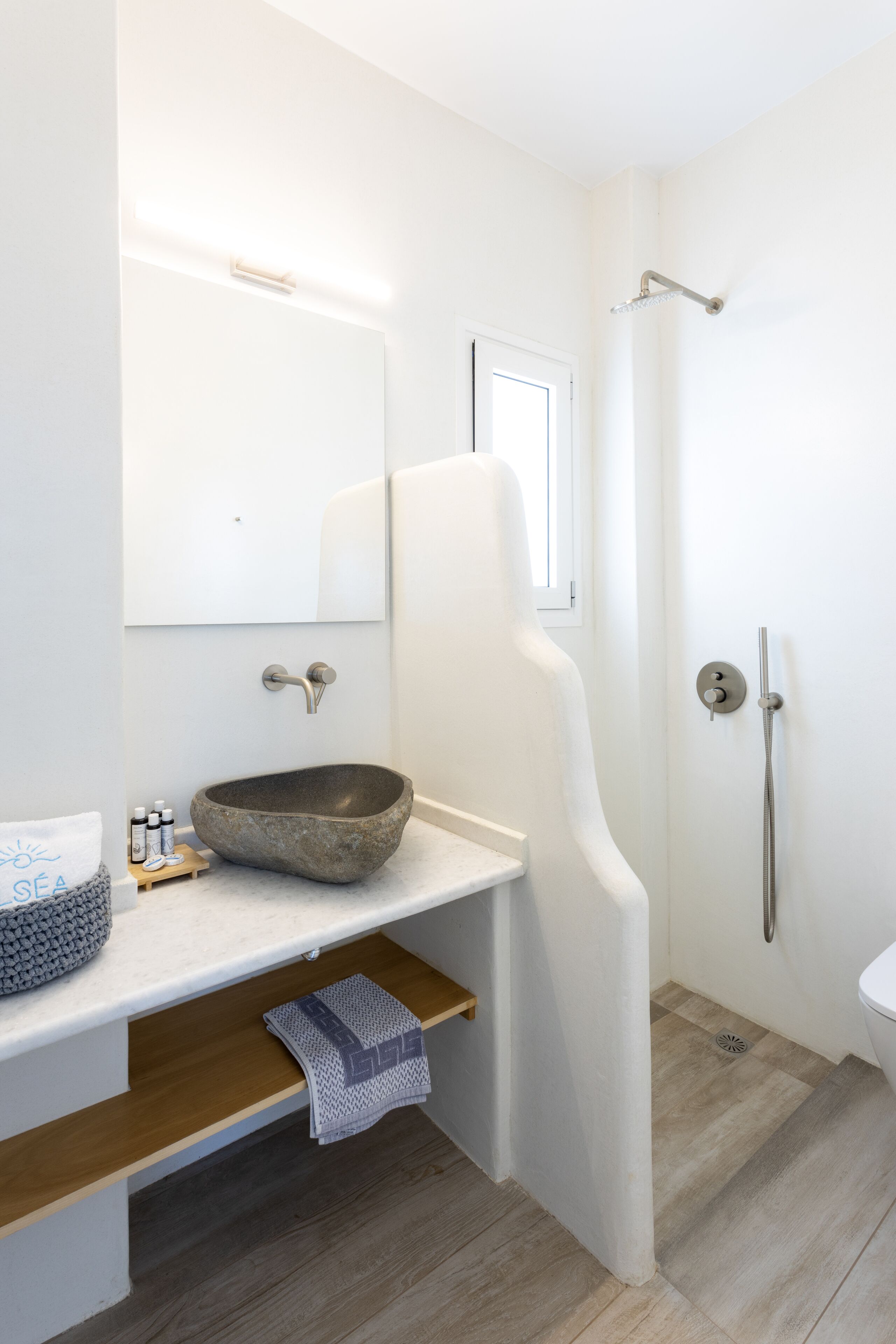 A modern bathroom with a stone basin, a mirror, a shower, and a wooden shelf with towels.