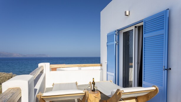 Un balcon avec deux chaises blanches dotées de coussins, une petite table en bois sur laquelle se trouvent une bouteille et un verre, et une porte bleue.