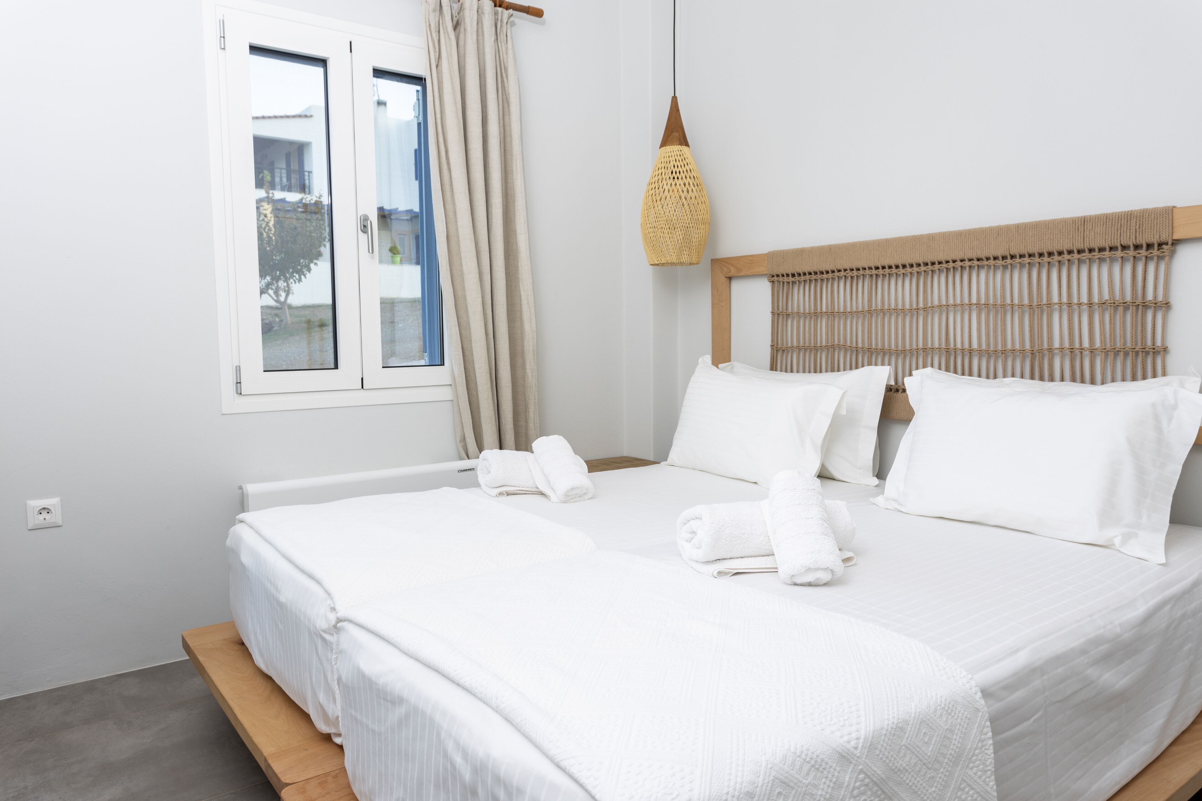 A neatly made bed with white linens and pillows, a wooden headboard, and a window with curtains.