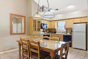 Une cuisine avec des meubles en bois, une table à manger avec des chaises, un four à micro-ondes, un réfrigérateur et des luminaires suspendus.