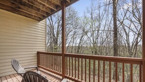 A covered porch with a wooden railing, two chairs, and a view of a wooded area.