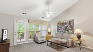 Un salon comprenant un canapé, un fauteuil, une table basse, deux lampes, un ventilateur de plafond et un tableau accroché au mur.