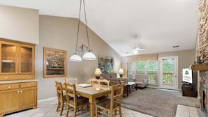 A dining area with a wooden table and chairs, a kitchen with wooden cabinets, a stone accent wall, and a living area with a sofa and lamp. - Golf Condo at StoneBridge only Minutes from Silver Dollar City (Reeds Spring)