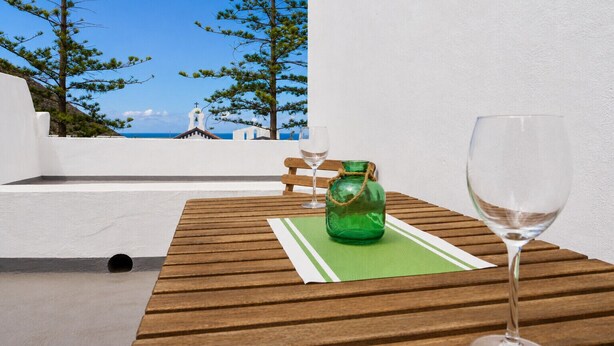 Une table en bois, sur laquelle sont posés un verre à vin et une bouteille verte, est placée devant un mur blanc, avec vue sur la mer et une église.