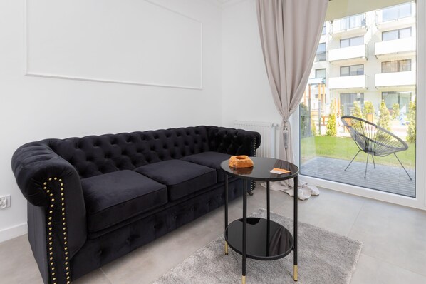 A modern living room with a black tufted sofa, a small round black table with a decorative item, and a view of a balcony with outdoor furniture.