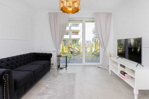 A modern living room with a black sectional sofa, a small round table, a white TV stand with books, and a large window with curtains. - Sunny Apartment with Terrace | Parking and Workspace (Gdynia)